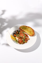 Sautéed shellfish medley with ciabatta toast, side view on white plate with leaf shadow art