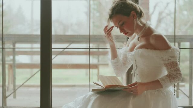 Young sexy woman reading paper novel book holding in hands sitting by the window. adult girl beauty face lips. have rest in royal room white vintage dress long ball gown old style blonde hair. 4k
