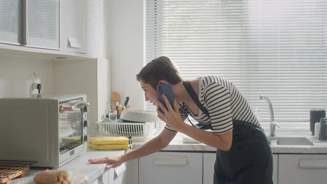 Medium long of Caucasian housewife in apron talking on mobile phone when cooking at home kitchen