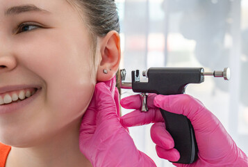 Cosmetic specialist in pink gloves pierces the ear of a smiling girl with a piercing gun.