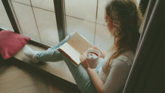 happy young woman holding cup of hot natural tea in hands drinking drink reading paper book. girl sits in cozy room at home near window sun light, cozy casual wear jeans t-shirt top back rear view 4k