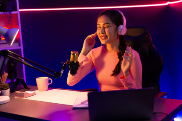 Fototapeta na wymiar Smiling creative young beautiful Asian influencer talking in channel with positive podcast speech at night time, using laptop for presentation online at pink neon light modern studio. Stratagem.