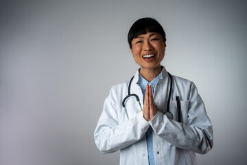 Cheerful female doctor with traditional Japanese greeting isolated on grey background.  Positive human face expressions, emotions, attitude. Portrait of a friendly, confident female healthcare doc