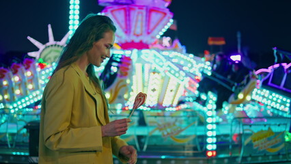 Romantic girl going illuminated luna park. Smiling woman holding sweet lollipop