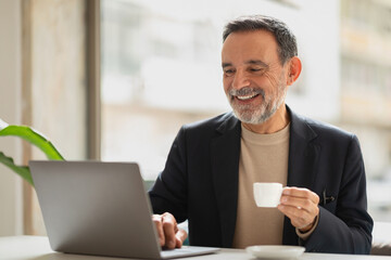 Cheerful senior man with a beard enjoying a cup of coffee while working on a laptop