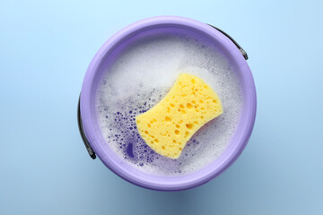 Bucket with foam and sponge on light blue background, top view