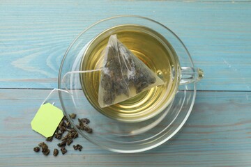 Tea bag in cup with hot drink and dry leaves on light blue wooden table, top view