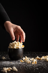 View of woman's hand over black bowl with popcorn on dark table with salt , selective focus, black...