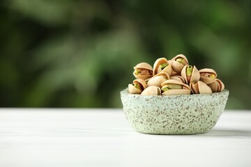 Tasty pistachios in bowl on white table against blurred background. Space for text