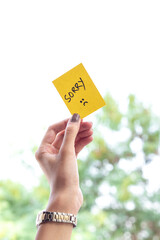 Young woman holding sticky paper note with sorry text and sad face emoticon over isolated background