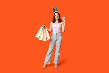 Beautiful young happy woman in bunny ears with shopping bags and credit cards on orange background....
