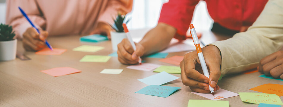 Group of young business group brainstorms ideas on colorful sticky notes. A portrait of startup group planing marketing strategy while writing down on sticky note at office meeting room. Variegated.