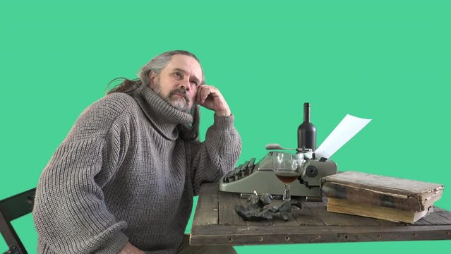 gray-haired mature male writer sits thinking about future works, then starts typing with one finger on an old typewriter - stands on a table with bottle of wine, blank letters and old books, green