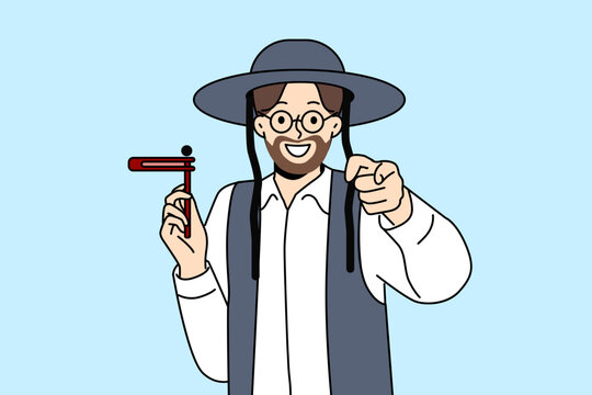 Orthodox jewish man with wooden grager ratchet in hand points finger at screen congratulating you on holiday of hanukkah. Israeli resident preaches judaism invites to celebrate hanukkah with smile