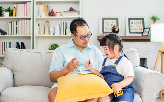 Asian father wearing casual clothes, sitting on sofa with little daughter girl, teaching homework, drawing, playing together with happiness in living room at home. Education, Relationship Concept.