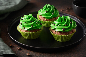 Three chocolate cupcakes with green whipped cream and chocolate sprinkles on dark brown background....