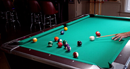 Green pool table in a bar, with a player striking the cue ball towards others. Motion blur of cue stick action.