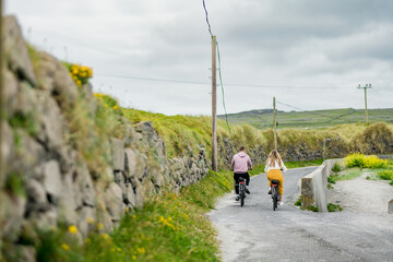 Tourists riding electric bikes on Inishmore, the largest of the Aran Islands in Galway Bay, Ireland. Renting a bicycle is one of the most popular way to get around Inis Mor. - obrazy, fototapety, plakaty