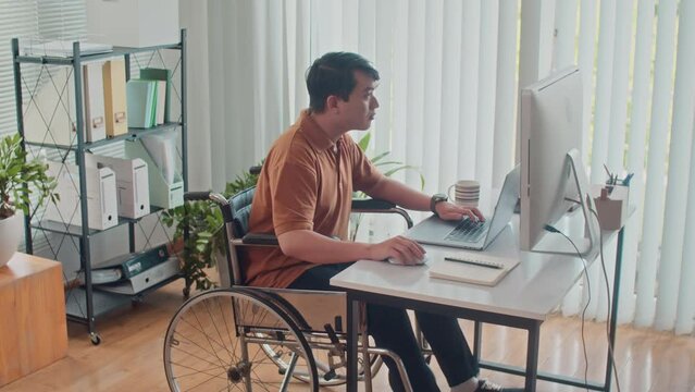 Long shot of pensive employee in wheelchair sitting in front of laptop while developing code on computer