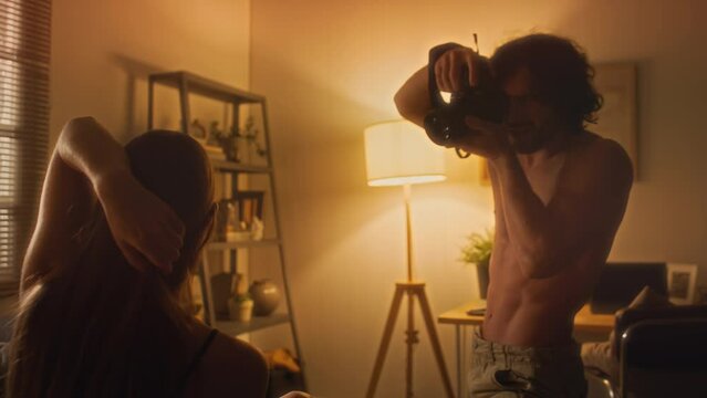Young shirtless man photographing girlfriend with digital camera as she posing on bed in cozy living room with warm light