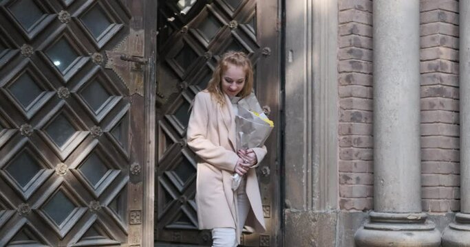 Joyful young blonde woman in beige coat walks out of building with massive wooden doors. Girl holds bouquet of flowers and smiles.
