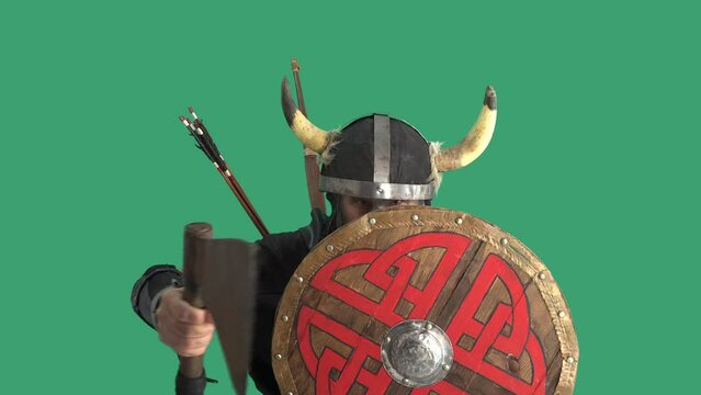 Portrait of scary Viking warrior, he threatens camera with large battle ax, screams loudly and knocks his weapon on wooden round shield. Green background