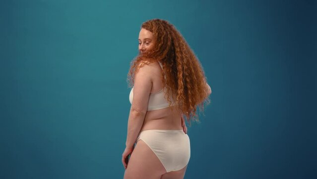 Young pretty redhead plus size or plump woman celebrating her natural body. Positive beautiful female model in white underwear against blue background. Body positive, skin care and cosmetology.