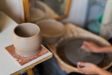 A newly shaped clay vase air-dries on a square tile beside a spinning pottery wheel being cleaned by a ceramist's hands, in a serene studio setting