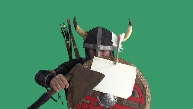 Portrait of mature Viking in helmet, warrior turns around and shows blank sheet of paper attached to an arrow on wooden shield. Aggressive looking, screaming and banging an ax on decorated shield