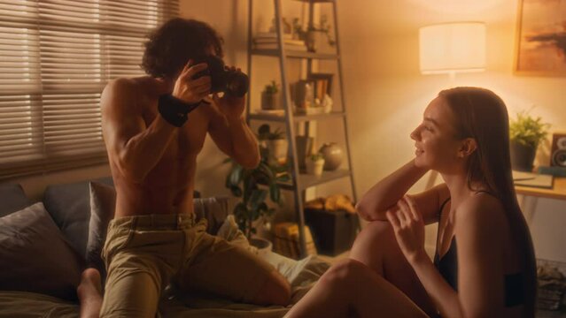 Man taking pictures with digital camera of his girlfriend in underwear smiling and posing on bed during home photoshoot in evening