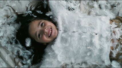 Joyful little girl lying in bed covered in feathers, carefree child looking at camera enveloped in...