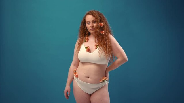 Young redhead plus size or plump woman with flowers celebrating her natural body. Positive beautiful female model in white underwear against blue background. Cosmetology, skin and body care concept.