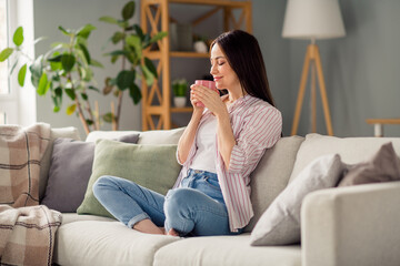 Full length photo of adorable peaceful person sit barefoot couch closed eyes smell fresh coffee mug weekend apartment inside