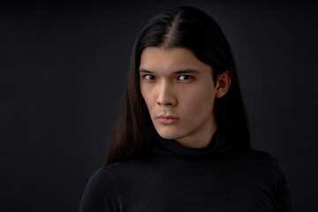 Fototapeta premium Portrait of confident handsome young man model with long straight hair in black shirt standing and looking at camera with serious proud face. indoor studio shot isolated on black background.