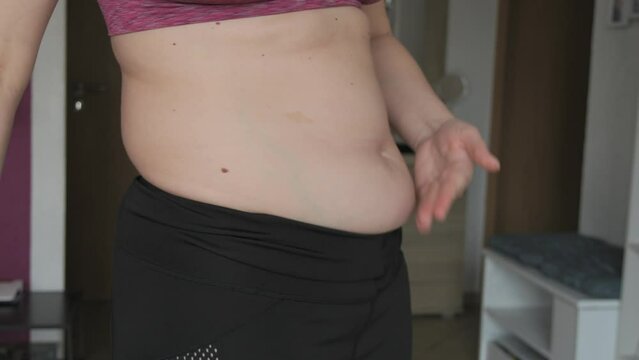 Female belly body positive. Woman touches belly folds in flat slow motion. A small layer of fat on abdomen of woman wearing a sports bra and leggings. Concept of sport, healthy lifestyle or overweight
