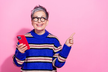 Photo of attractive pensioner woman hold device point look empty space wear trendy blue striped clothes isolated on pink color background