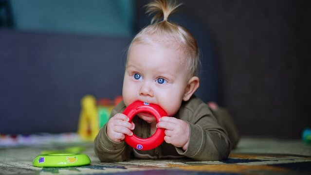 Cute blue-eyed baby lies on the floor in the room. Beautiful child is chewing his toy ring. Close up.