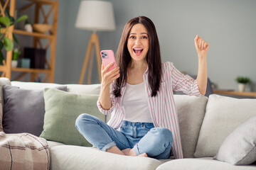 Obraz premium Full size photo of delighted crazy girl sitting couch use smart phone raise fist achievement modern apartment indoors