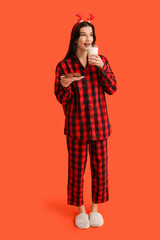 Beautiful young woman in checkered pajamas with sweet cookies and glass of milk on orange background
