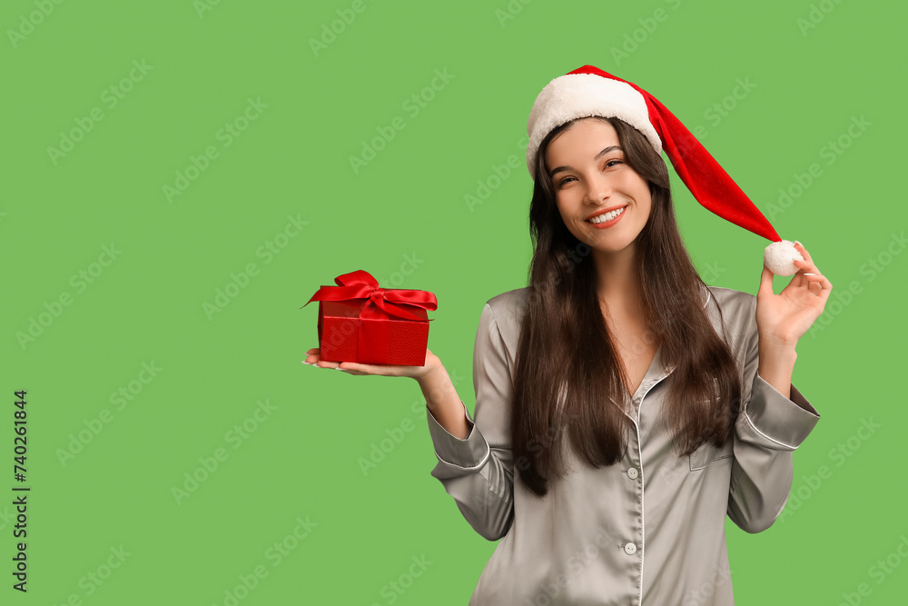 Sticker beautiful young woman in grey pajamas and santa hat with christmas gift box on green background