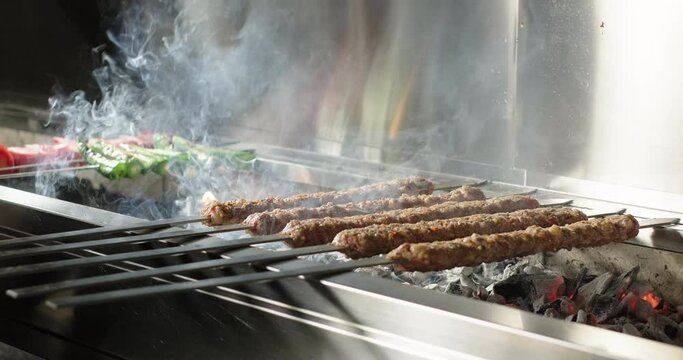 Adana kebab barbecue on charcoal grill in a restaurant. Smoke rising over traditional Turkish kebab, tomatoes and peppers grilled on skewers. Close-up