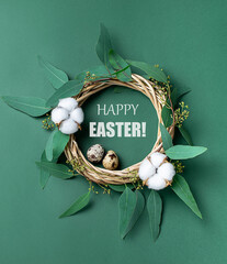 Beautiful festive composition with straw wreath decorated with eucalyptus leaves, cotton flowers, eggs and text Happy Easter on green pastel background. Eco friendly zero waste festive decorations.