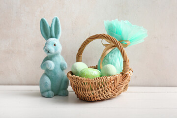 Fototapeta na wymiar Wicker basket with painted Easter eggs, toy bunny and gift bag on white wooden table