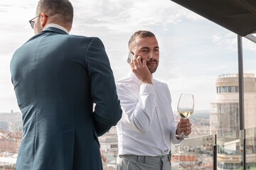 Two men in suits with wine while one of them talks on the phone