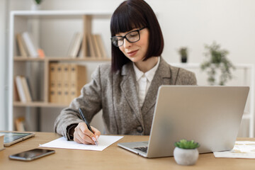 Fototapeta premium Mindful woman in eyeglasses making notes with pen while looking at laptop screen in workplace. Efficient Caucasian employee doing online research on company's development using wireless technologies.