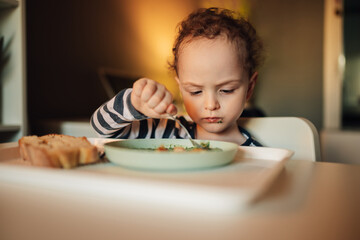 An adorable child is using spoon for eating his healthy lunch at home.