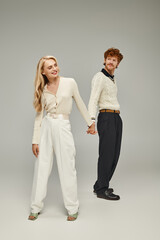 full length of happy young redhead man and blonde woman holding hands on grey backdrop in studio