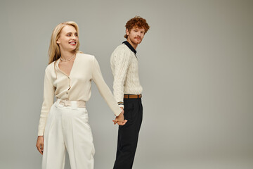 happy blonde woman and redhead man in trendy casual clothes holding hands on grey, old fashion style