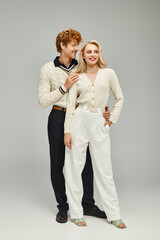 full length of smiling redhead man in casual clothes embracing blonde woman on grey, trendy couple