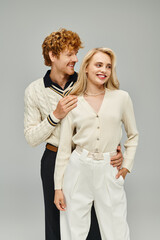 happy redhead man in casual attire embracing young blonde woman on grey backdrop, fashionable couple
